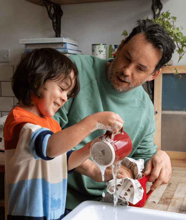 A waist-up shot of a male child and his dad washing and drying mugs in the sink together at home.