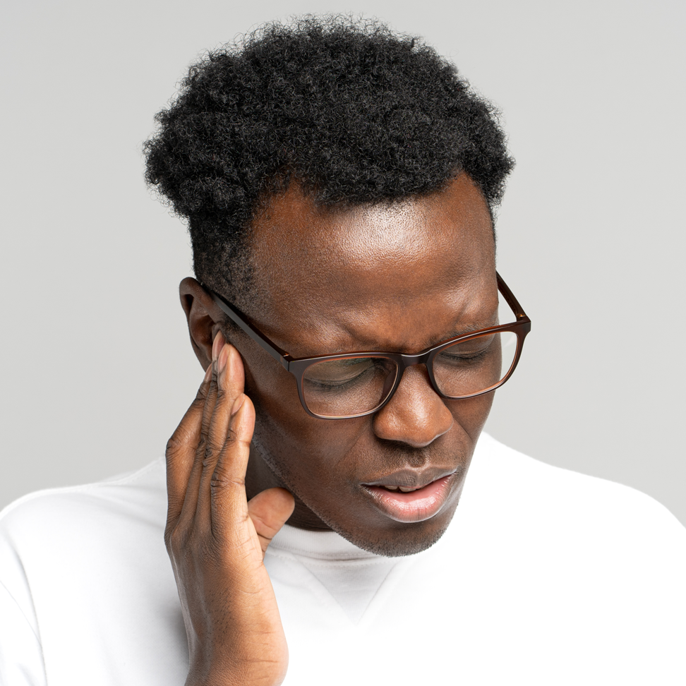 Portrait of young man wearing glasses against a grey background. He is holding his ear and appears to be in pain.